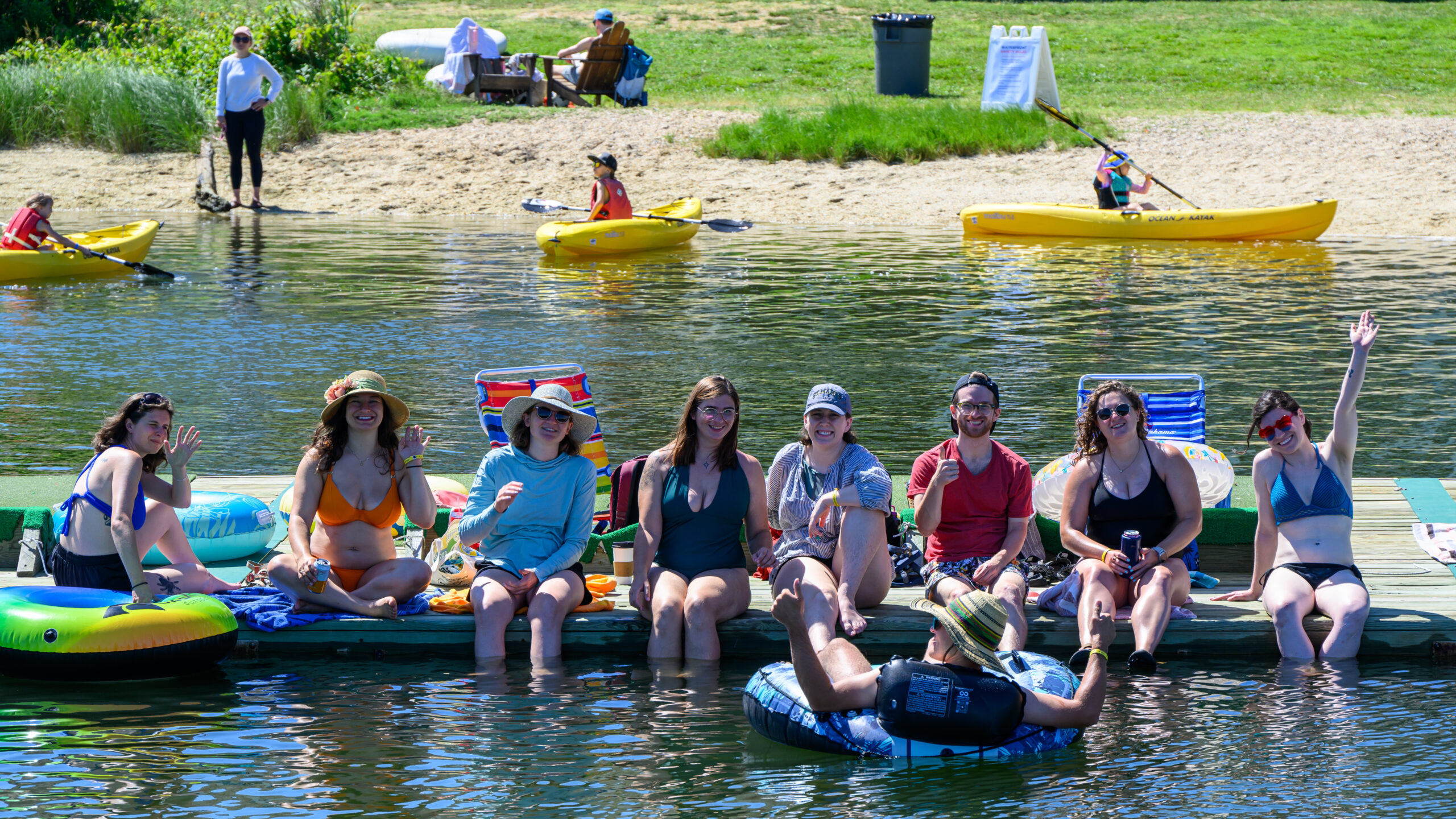 A group of people in swimsuits sit on a dock by a lake, some holding drinks and waving, with kayakers and others relaxing in the background on a sunny day.