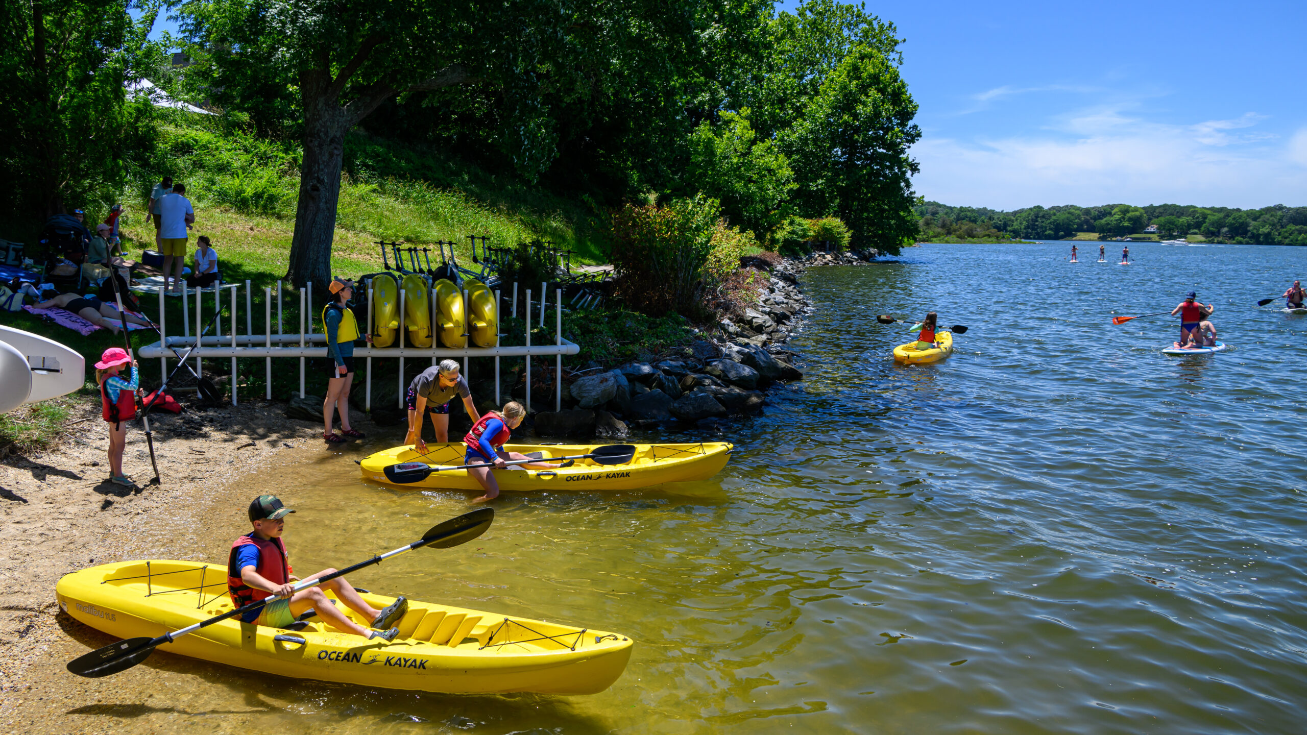 People prepare yellow kayaks on a sandy lakeshore, while others paddle on the water. Kayaks are stacked nearby and trees provide shade along the edge of the lake.