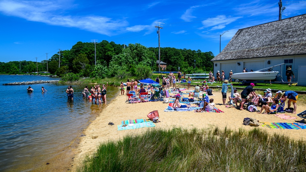 People relax on towels and swim at a small sandy beach by the water, with a building, grassy area, and trees in the background under a clear blue sky.
