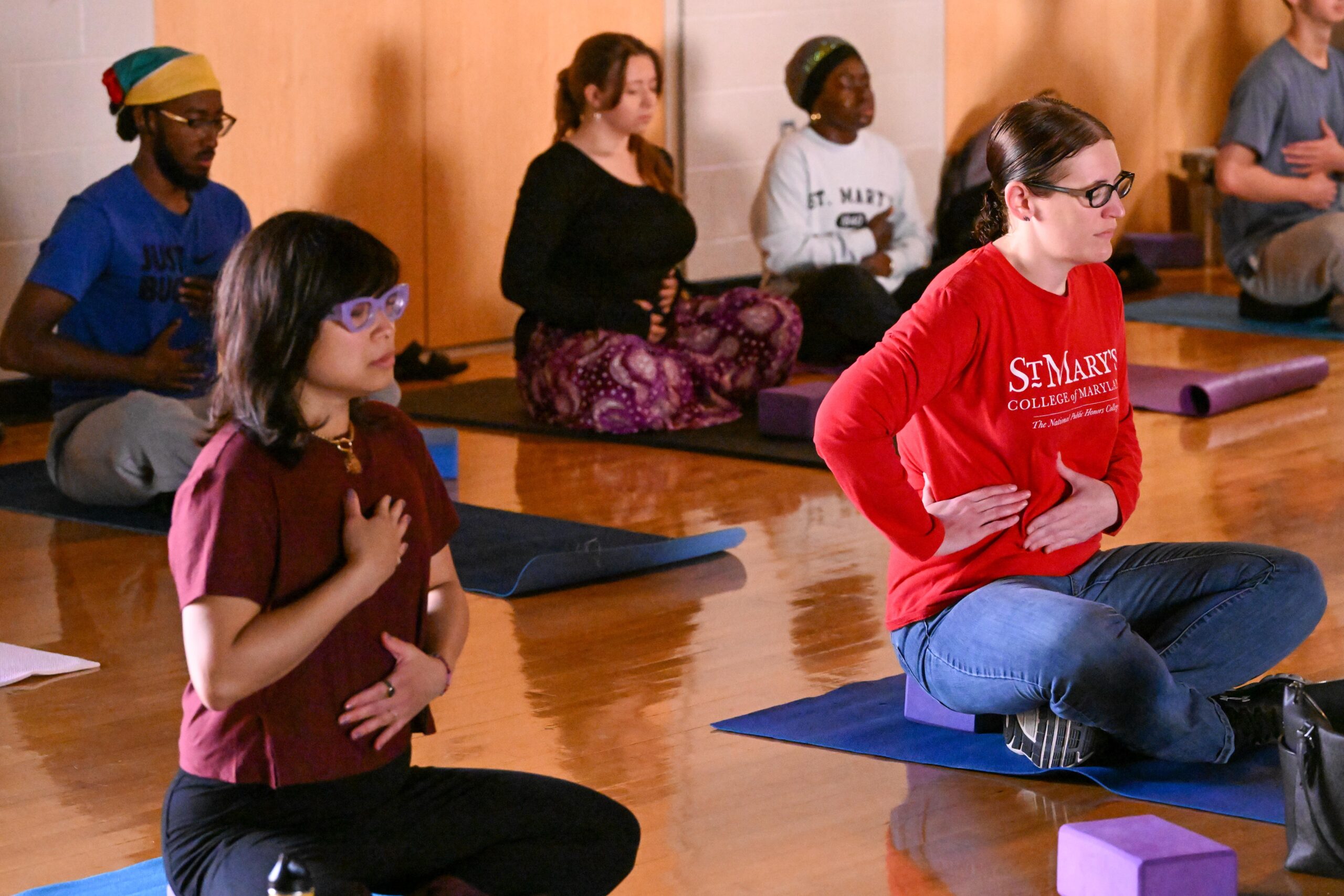 A group of people sit cross-legged on yoga mats in a studio, practicing deep breathing exercises with hands placed on their chests and abdomens.