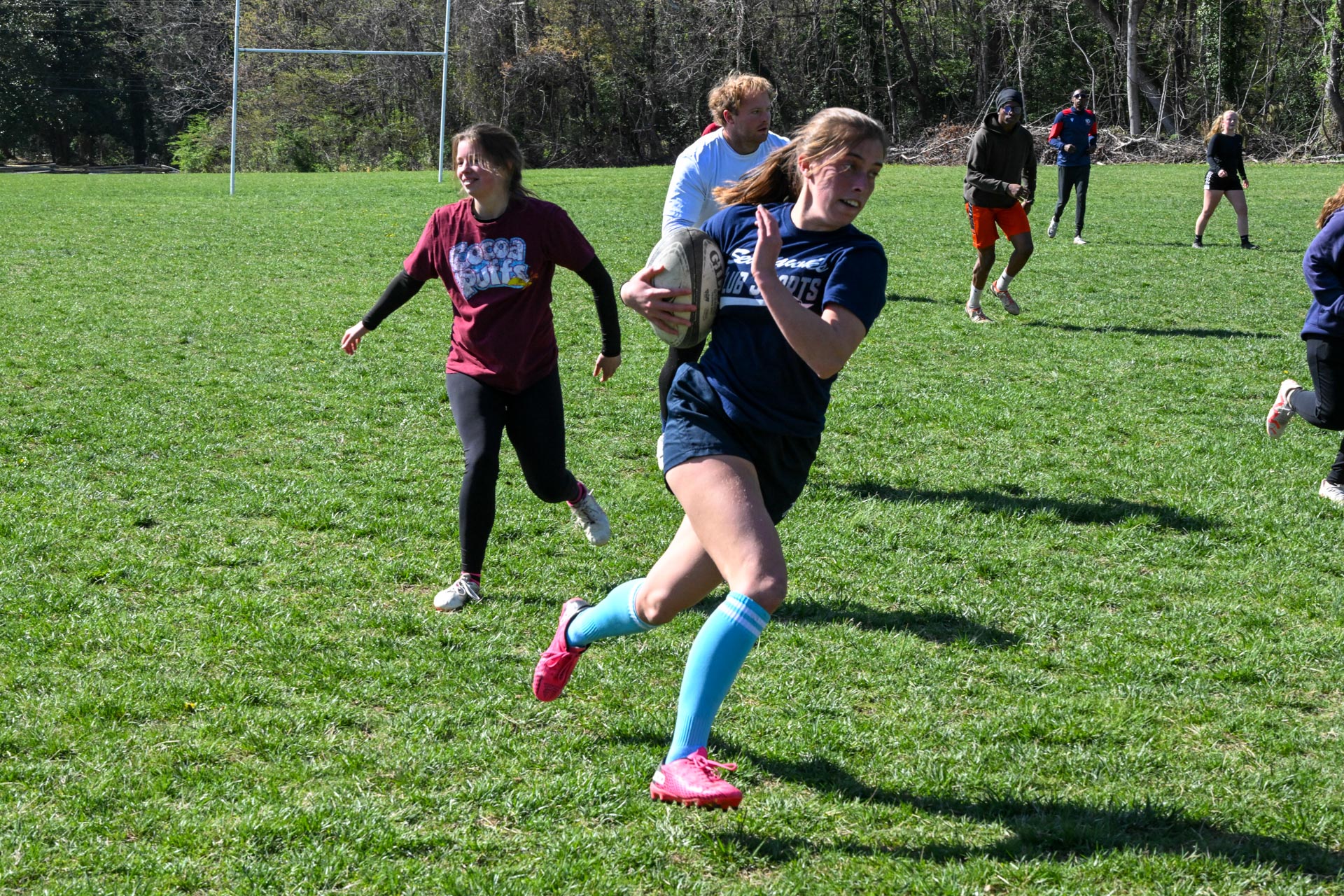 A woman runs with a rugby ball on a grassy field while other players chase her during a game.