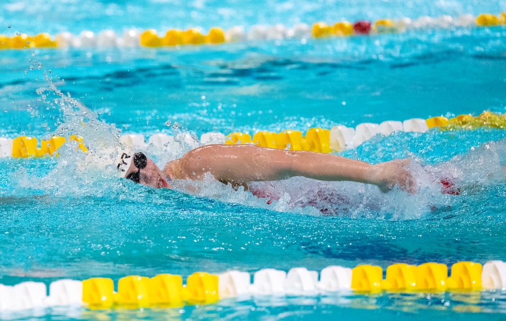 A swimmer wearing goggles and a swim cap performs freestyle in a lane of an indoor pool, surrounded by yellow and white lane dividers.