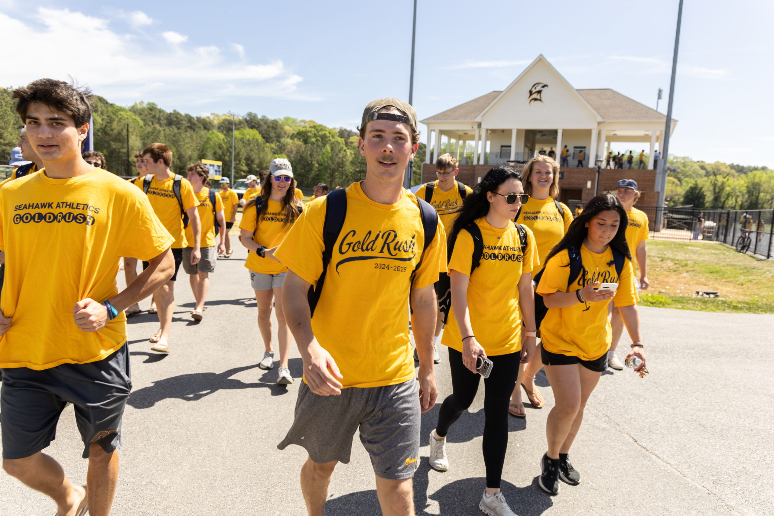 A group of students in yellow "Gold Rush" shirts walk together outdoors near a sports field and a building with a porch.