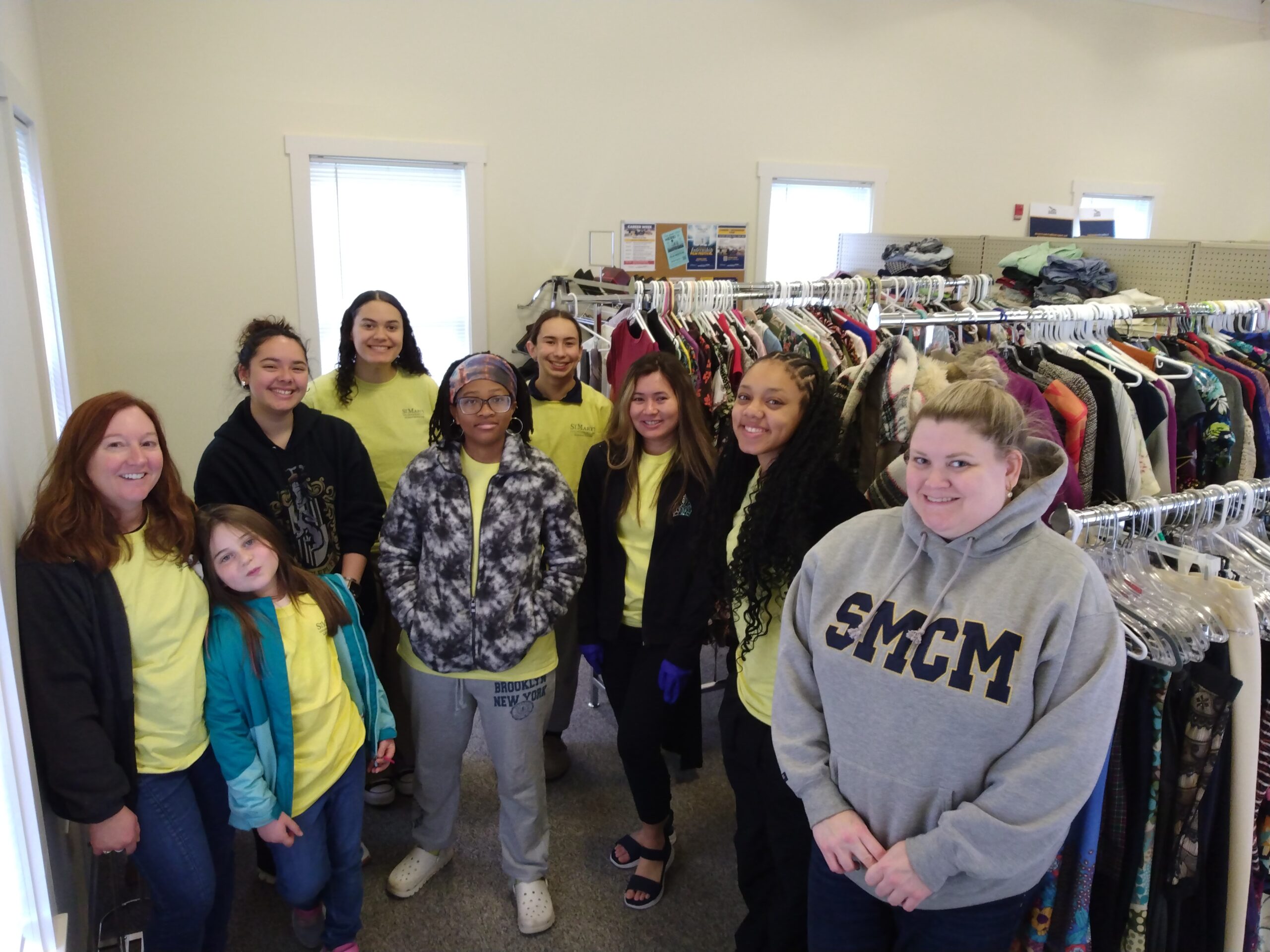 Nine people, mostly women and girls, stand and smile in a small thrift store, with clothing racks and folded clothes visible in the background.