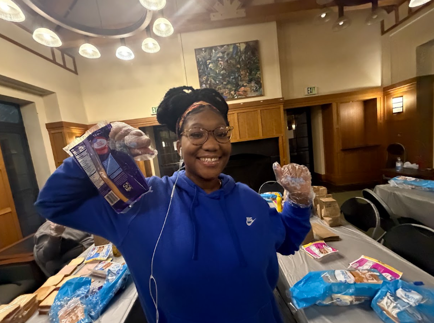 A person wearing a blue hoodie and gloves smiles while holding food packaging in a room set up for meal prep, with tables covered in supplies.