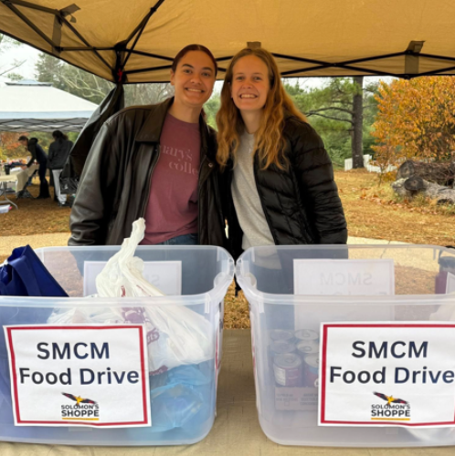 Two people stand under a canopy behind bins labeled "SMCM Food Drive," collecting food donations outdoors on a cloudy day.
