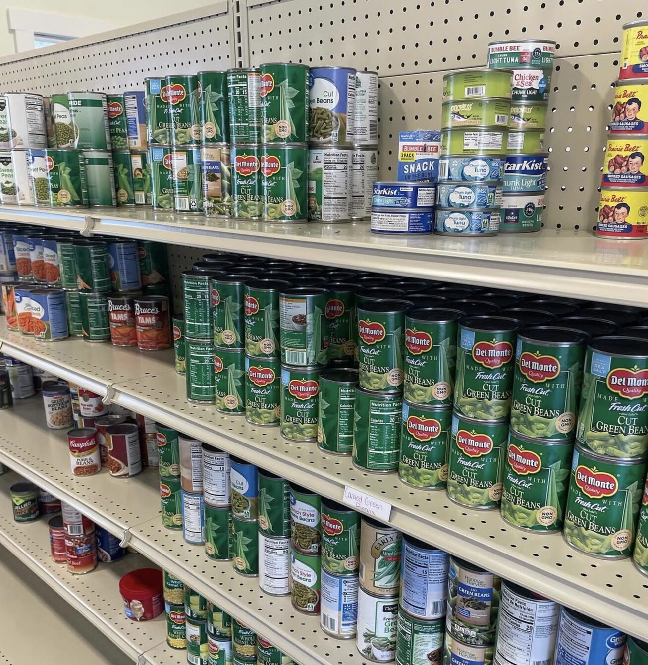 Shelves in a store display canned vegetables, beans, fruit, and tuna. Most cans are green-labeled Del Monte brand, arranged in rows on beige shelves with pegboard backing.