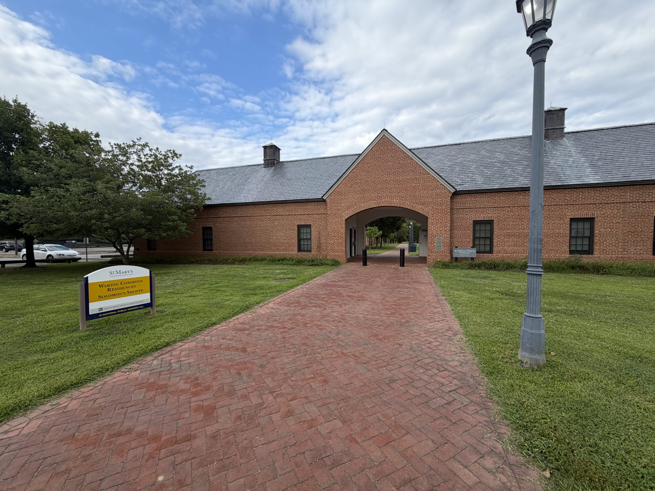 A brick walkway leads to an arched entrance of a red brick building with a lamp post and a yellow campus sign on the left.