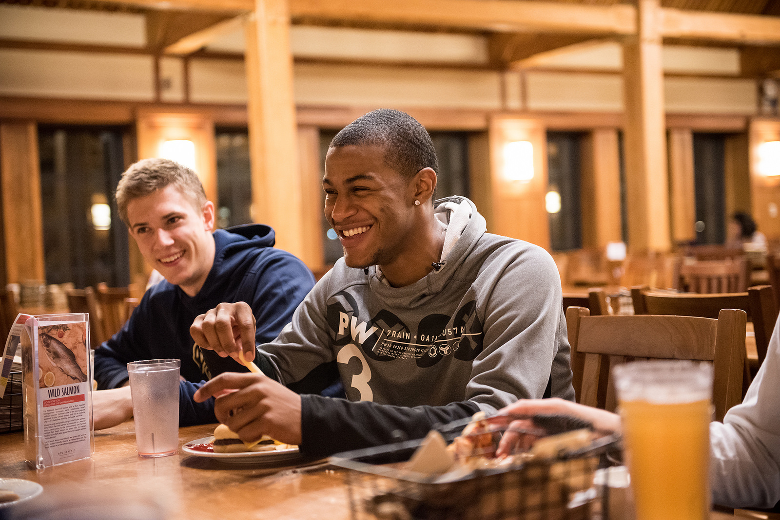 Two young men sit at a wooden table in a restaurant, smiling and enjoying their dining experience as they eat. A menu and drinks are visible on the table in front of them.