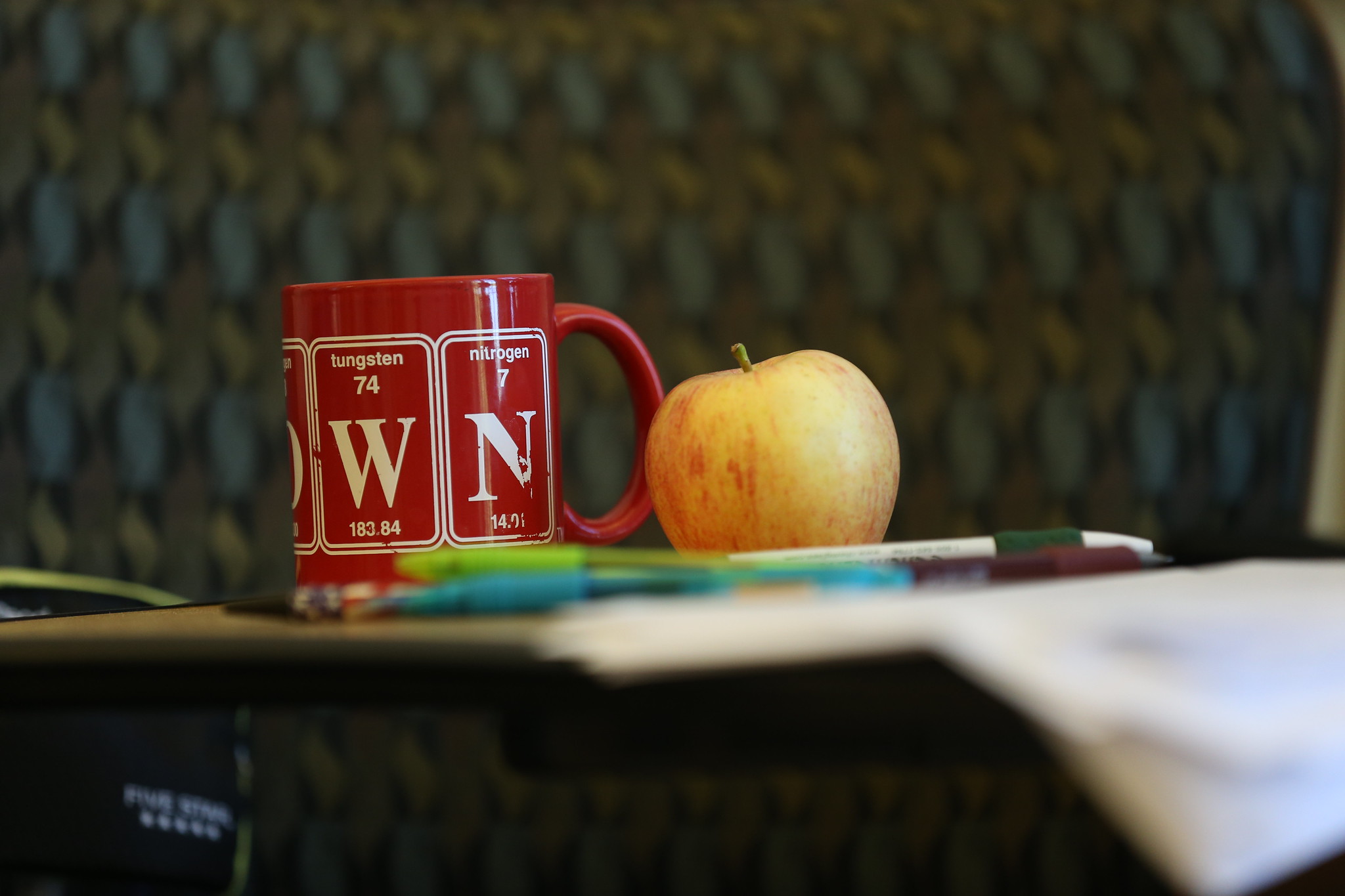 A red mug with periodic table symbols, a yellow-red apple, and some pens are on a desk. A blurred patterned chair serves as the backdrop.