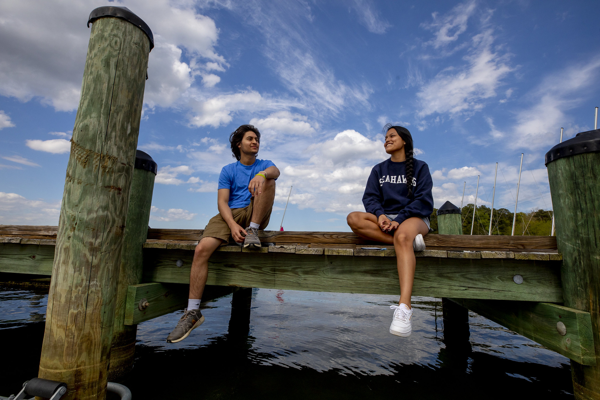 Two people sit on a wooden dock over water, chatting and smiling. One wears a blue shirt and khaki shorts, and the other wears a navy sweatshirt and white sneakers. A bright sky with clouds is overhead, and sailboats are in the background.
