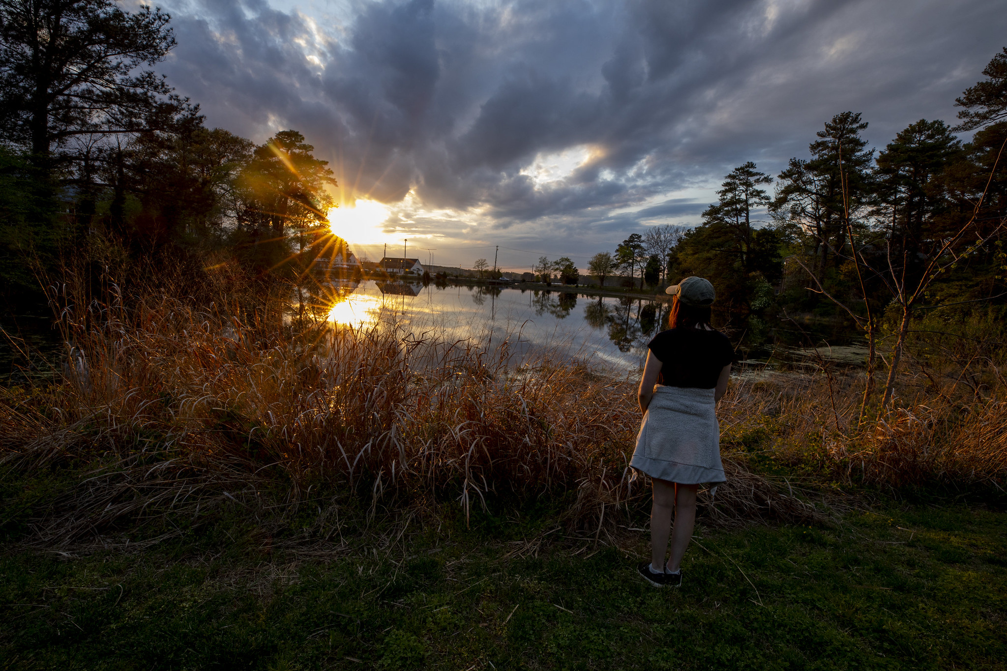 A person stands on a grassy area by a lake, gazing at a dramatic sunset with clouds scattered across the sky. Tall grasses and trees surround the lake, while the sun casts reflections on the water.