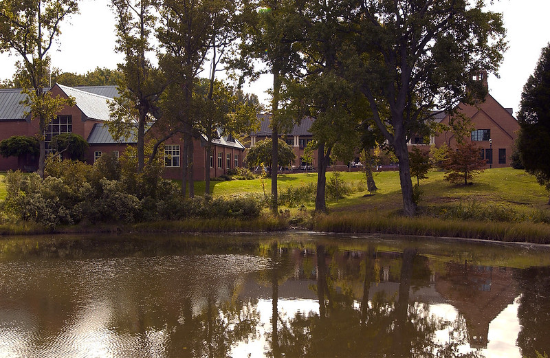 A serene pond reflects surrounding foliage and large brick buildings. Trees line the grassy bank, partially obscuring the buildings. The sky is clear, and sunlight illuminates the scene, creating a peaceful atmosphere.