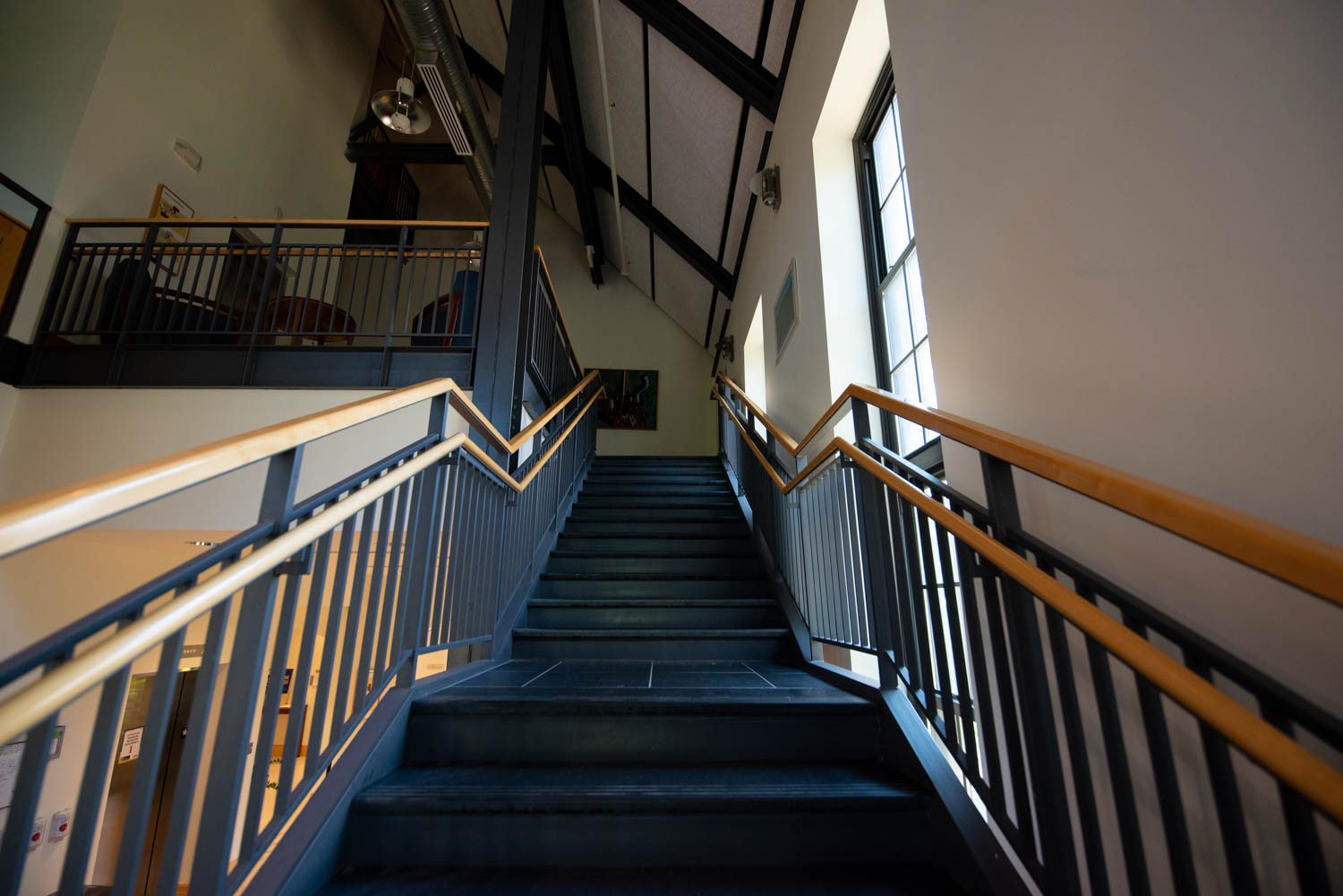 A well-lit, wide staircase leads upwards in a modern building. It has black steps and wooden handrails. Large windows on the right allow natural light to fill the space, illuminating the gray walls and high ceiling.