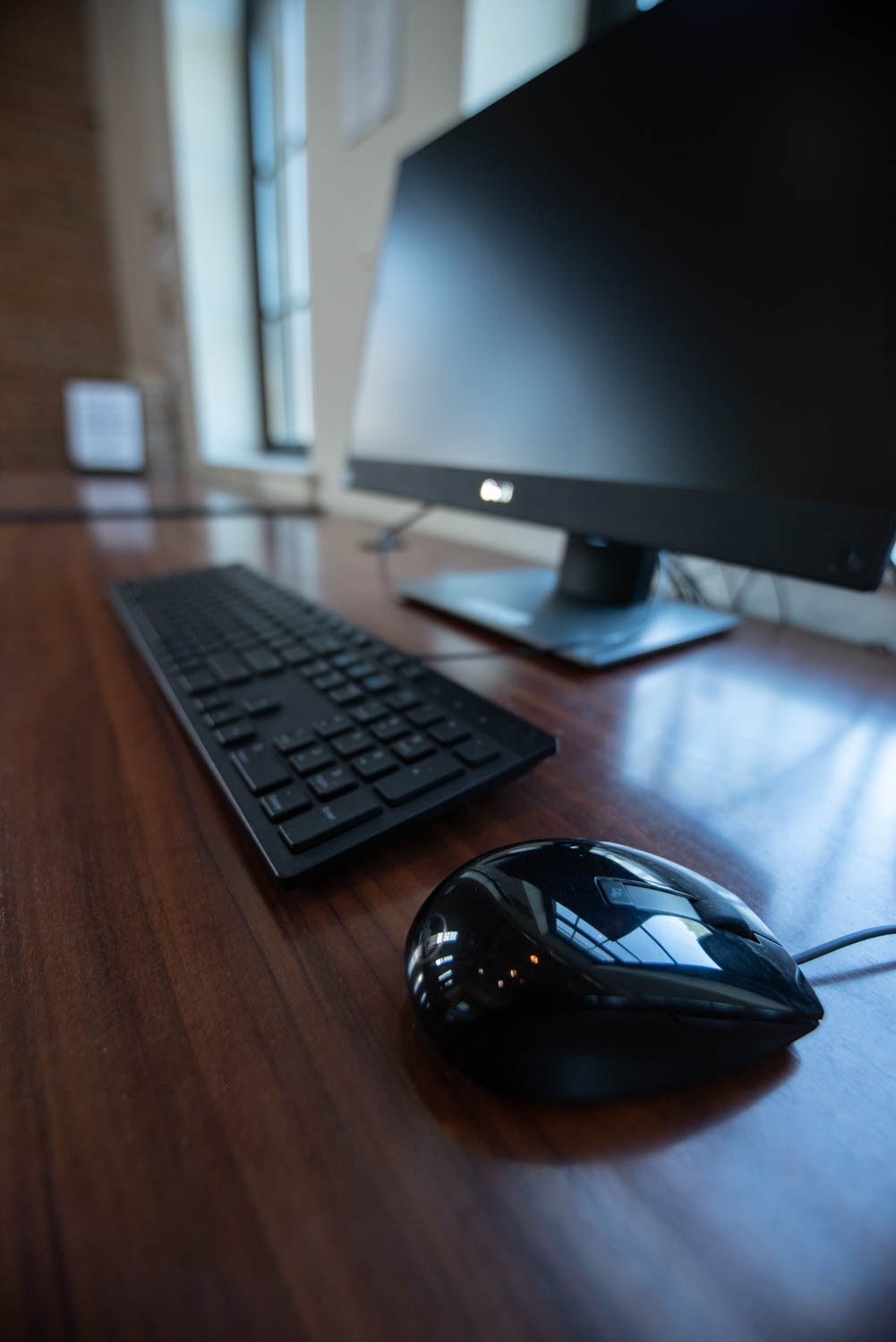 A sleek black computer monitor on a wooden desk, accompanied by a matching keyboard and mouse. Soft natural light filters through a window, reflecting on the mouse surface.