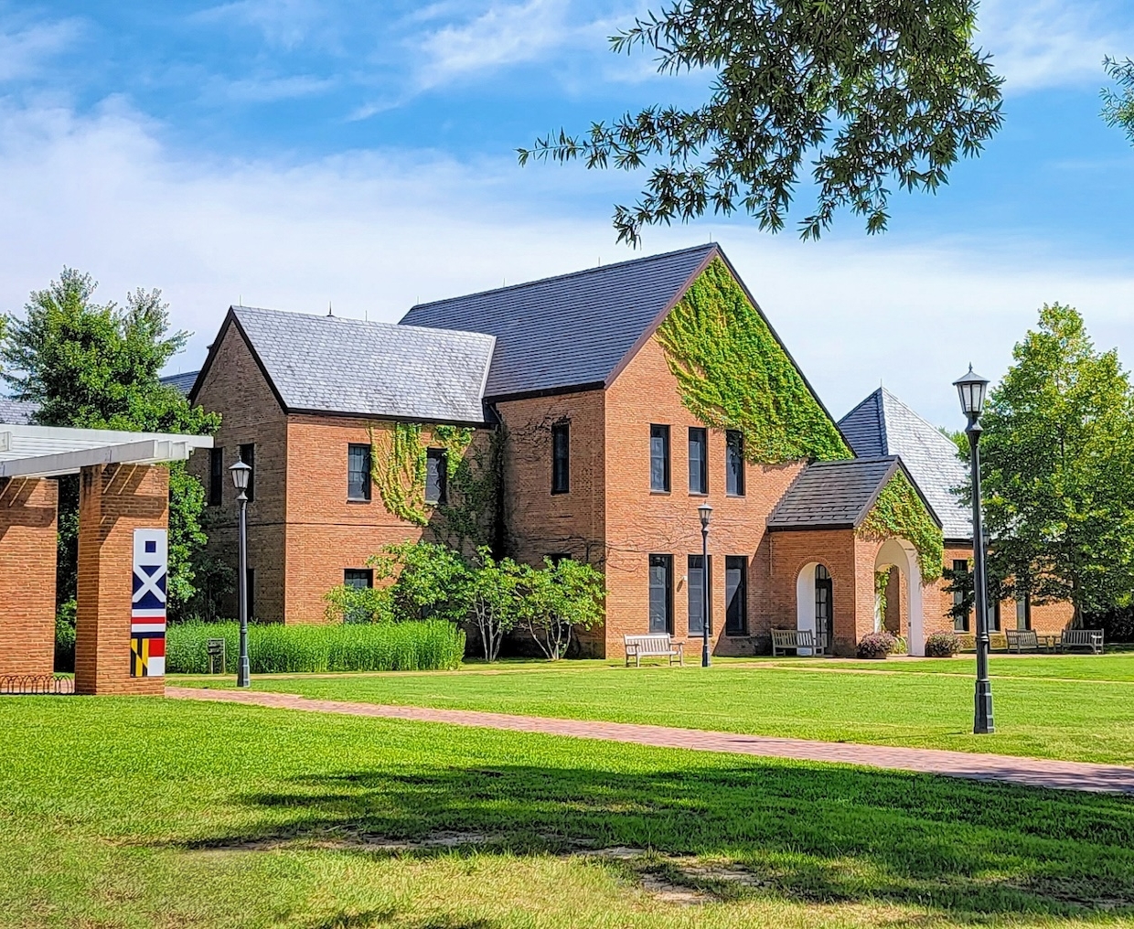A brick building with a slate roof is partially covered in ivy. It sits on a large grassy area with trees. A paved path leads to it from the front. The sky is blue with a few clouds.