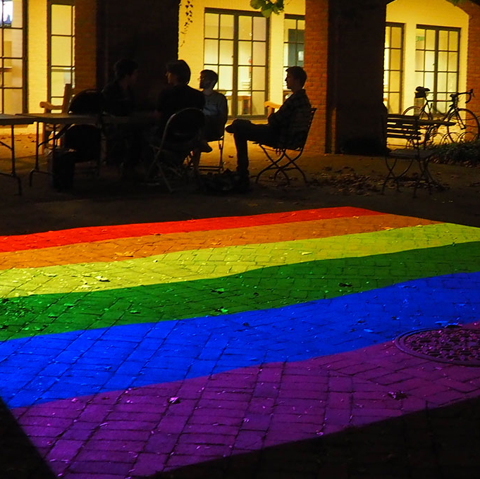 A rainbow flag is projected onto a brick pavement at night, with people sitting and talking at a table in the dimly lit background.