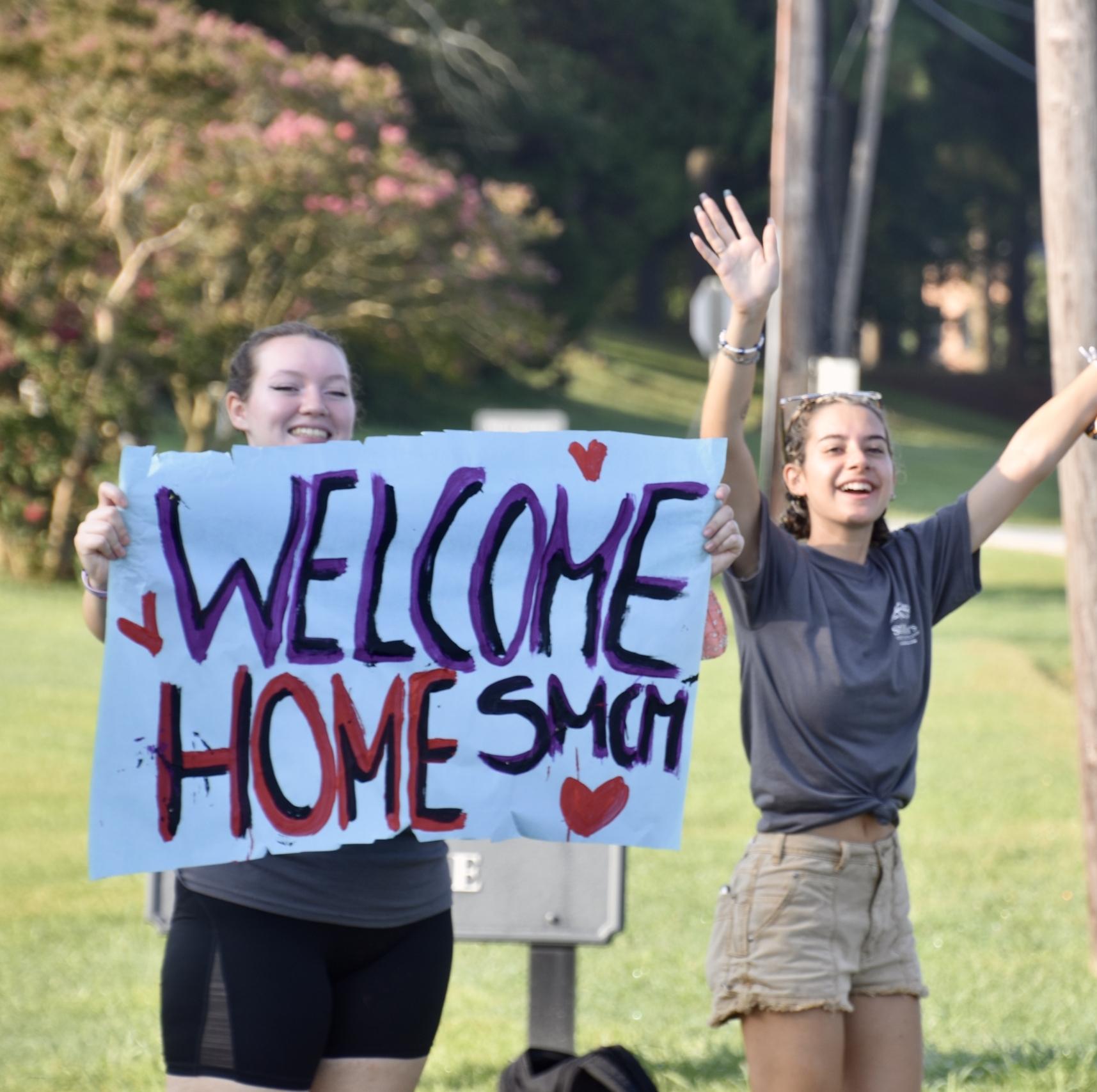 Two smiling individuals stand outside on grass, holding a sign that reads "Welcome Home SMCM" with hearts. One person has both arms raised, and both are dressed casually. Trees and a road are visible in the background.