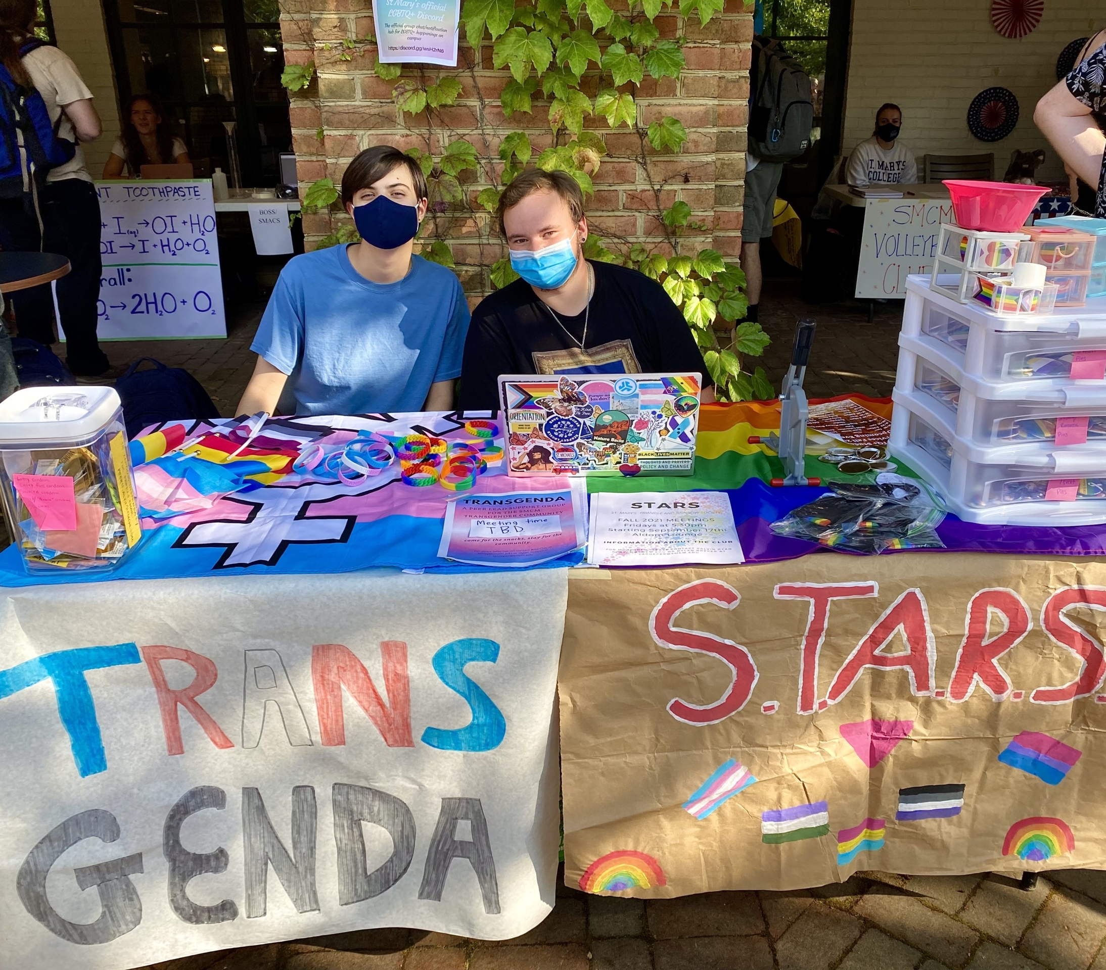 Two people sit at a booth with colorful banners reading "TRANSGENDA" and "S.T.A.R.S." The table is covered with rainbow flags and various informational materials. Both individuals wear masks and smile at the camera.