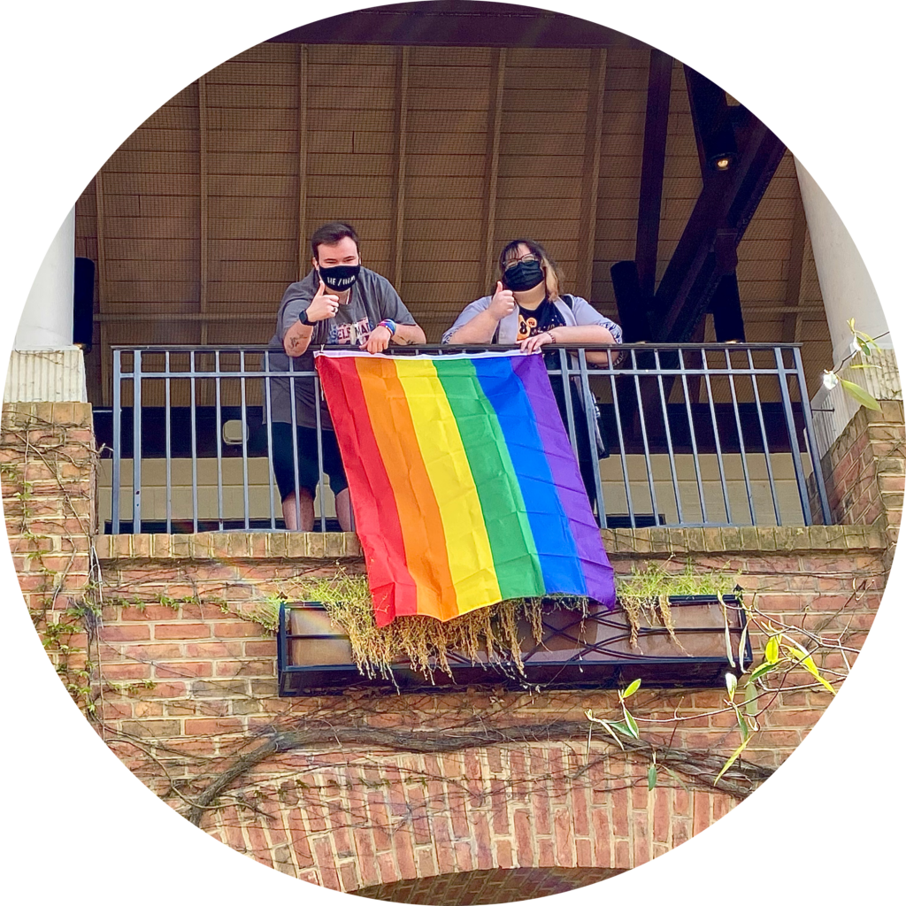 Two people wearing masks stand on a balcony, leaning on a metal railing. A rainbow flag hangs over the railing. The balcony is part of a brick building with vines and white pillars, and they give thumbs-up gestures.