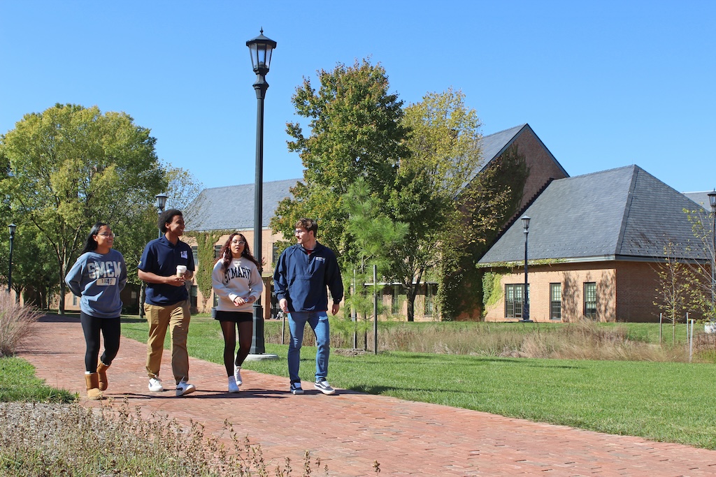Four students walk together on a brick path in a sunny campus setting, surrounded by grass and trees. They wear casual clothing and carry books. Buildings with sloped roofs are visible in the background under a clear blue sky.