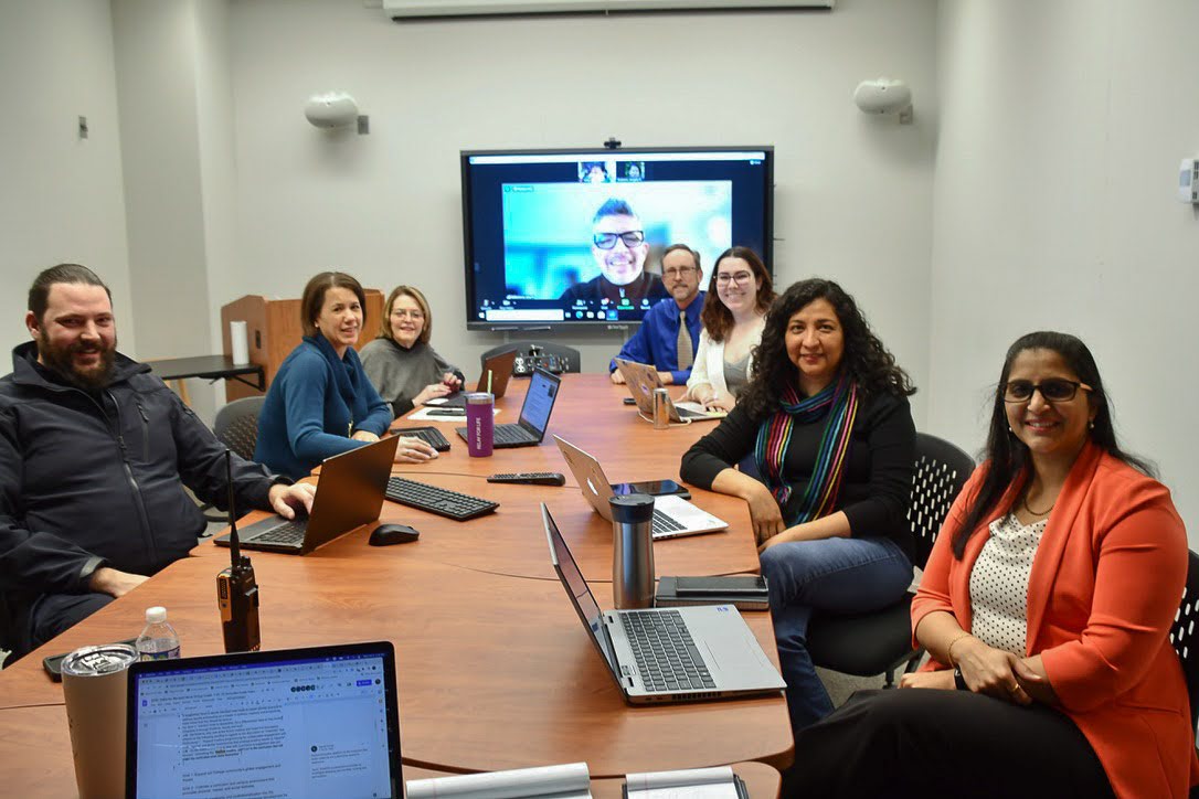 A group of eight people, seven in a room and one on a screen, are seated around a conference table with laptops and notebooks. They are smiling and appear to be engaged in a meeting or discussion.