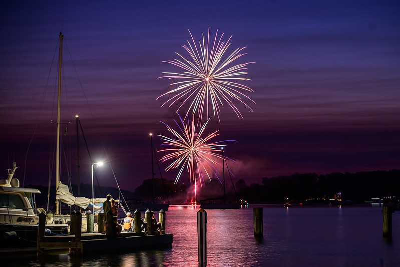 Fireworks light up the evening sky over a calm waterfront with boats docked and people standing on the pier.