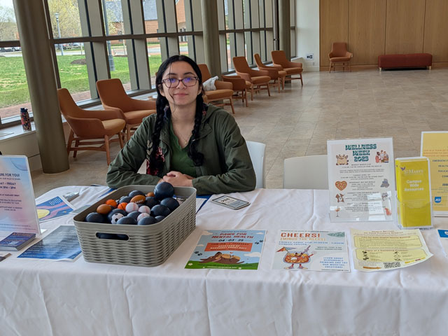 A person sits behind a table with wellness information, pamphlets, and a basket of stress balls in a bright, modern lobby area.