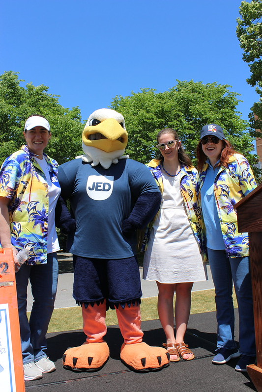 Four people, including a person in an eagle mascot costume with a "JED" shirt, stand outdoors posing for a photo on a sunny day.