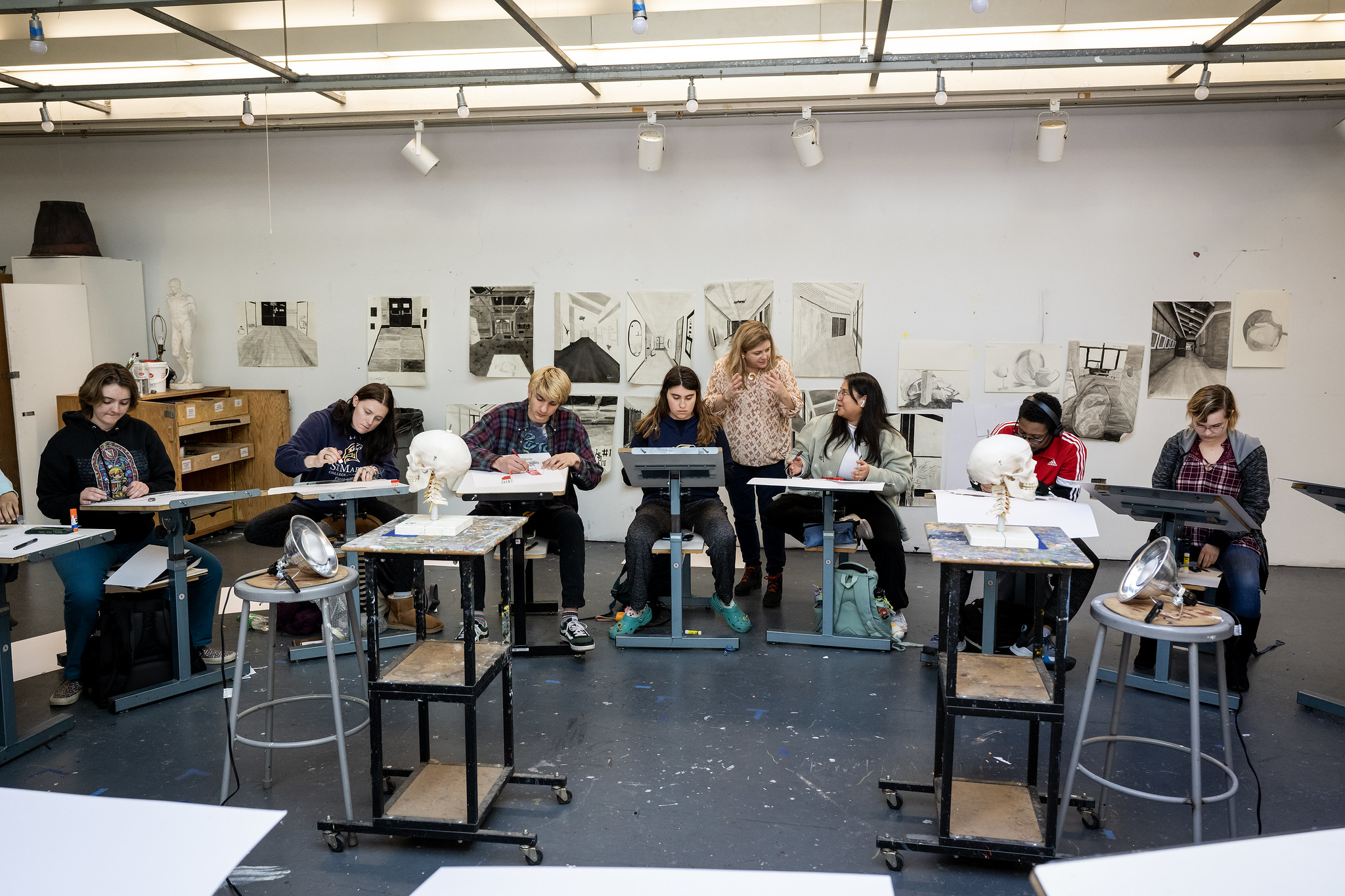 A group of students in an art classroom sketching plaster busts at easels, with drawings displayed on the back wall.