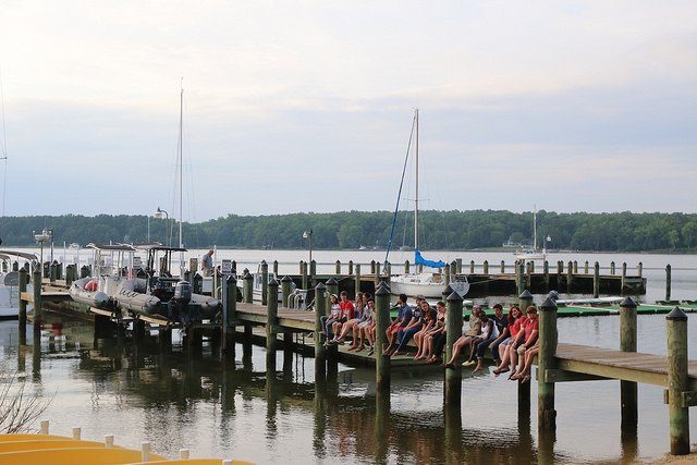 A group of people, mostly wearing red and blue, sit along the edge of a wooden dock extending over calm water. Boats are moored nearby, and trees are visible in the background under a lightly clouded sky.
