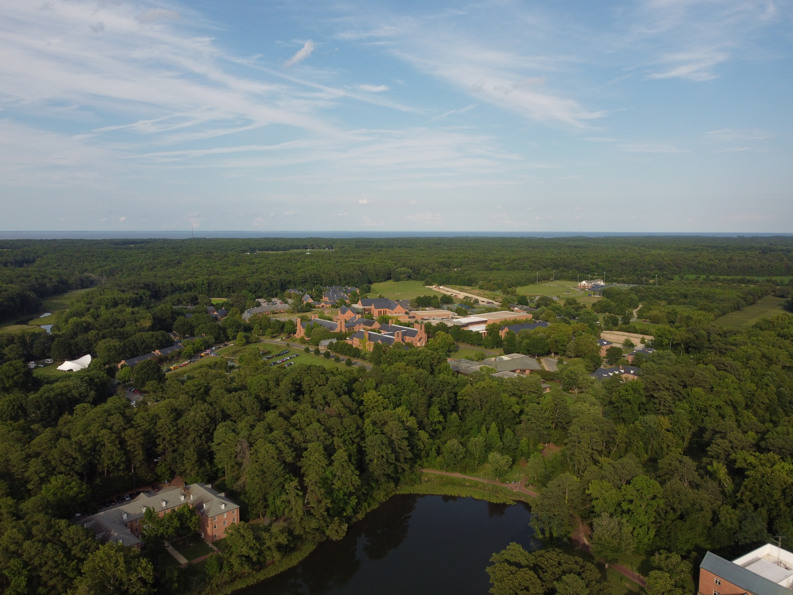 Aerial view of a campus with multiple brick buildings surrounded by dense trees, a pond in the foreground, and open sky above.