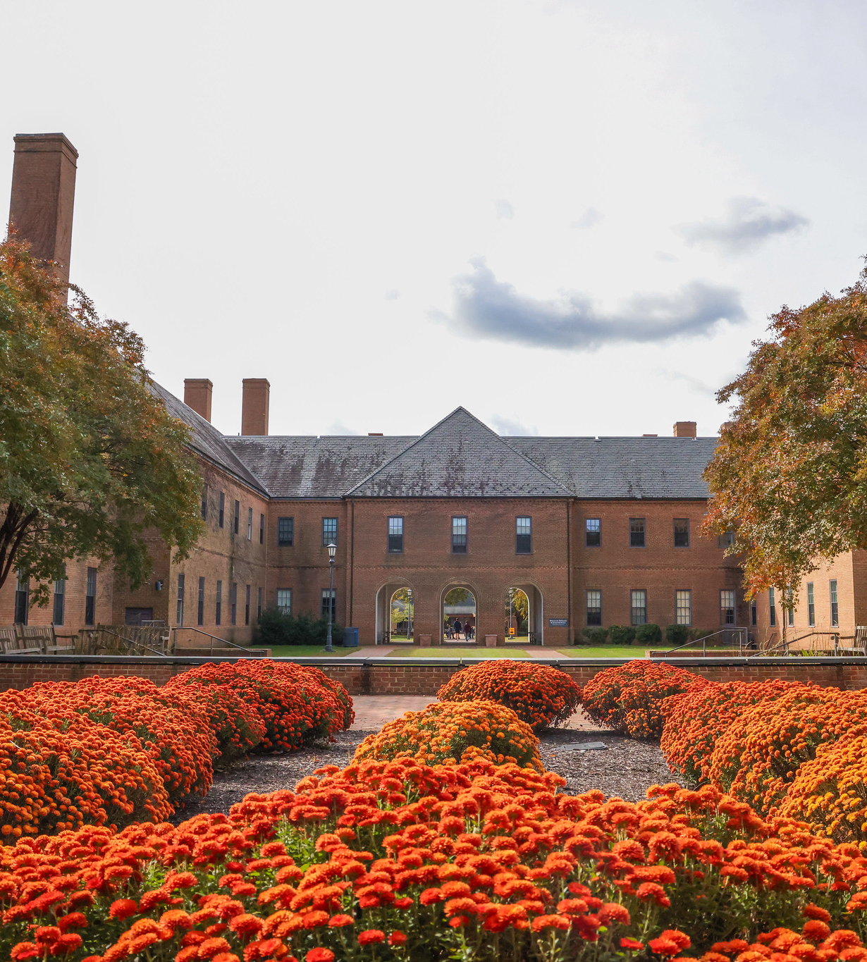 A large brick building with tall chimneys is framed by trees and flower beds with bright orange blooms in the foreground.