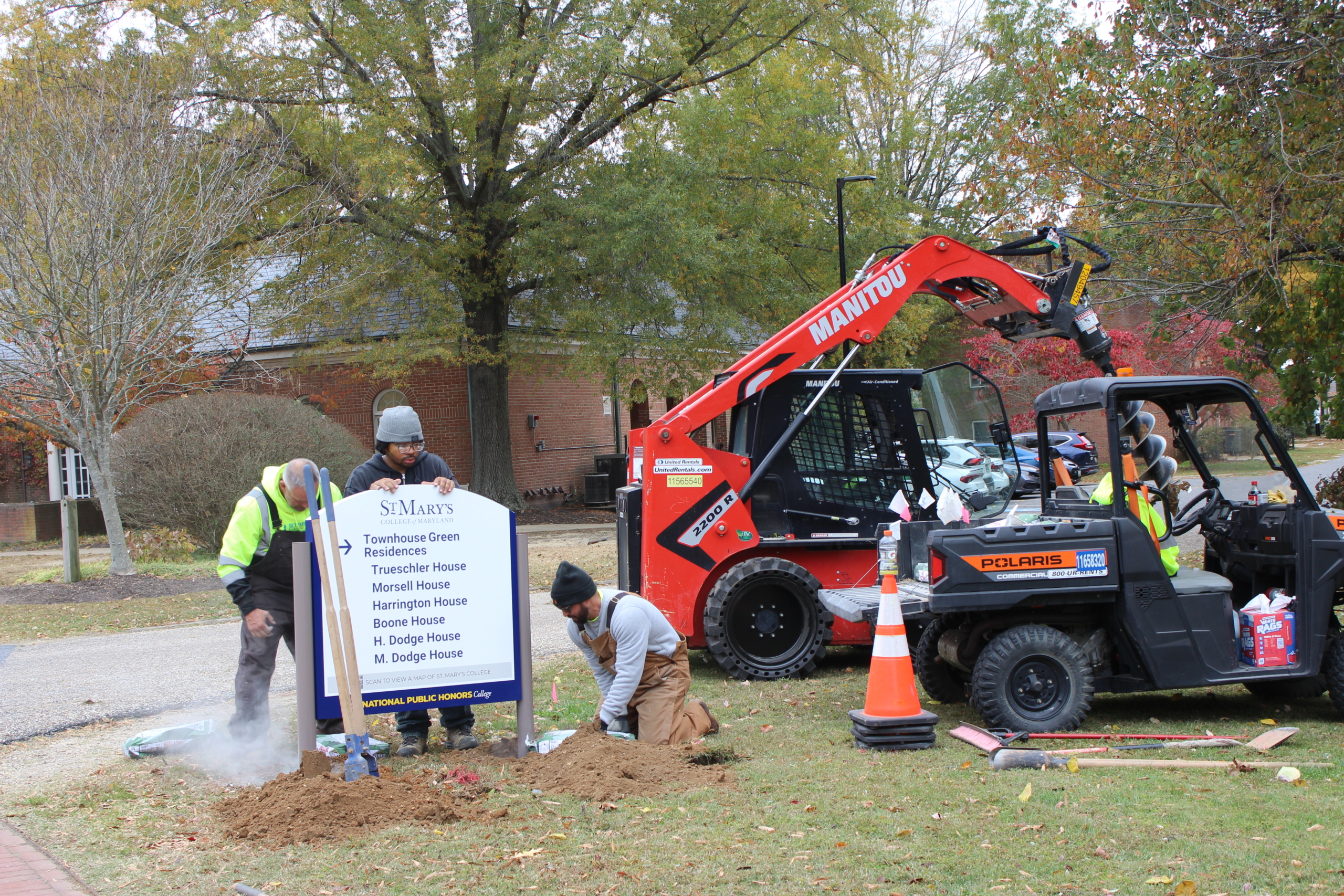 Three workers install a St. Mary’s sign on a lawn near a brick building, using tools and vehicles including a red forklift and a black utility cart. Orange cones mark the work area.