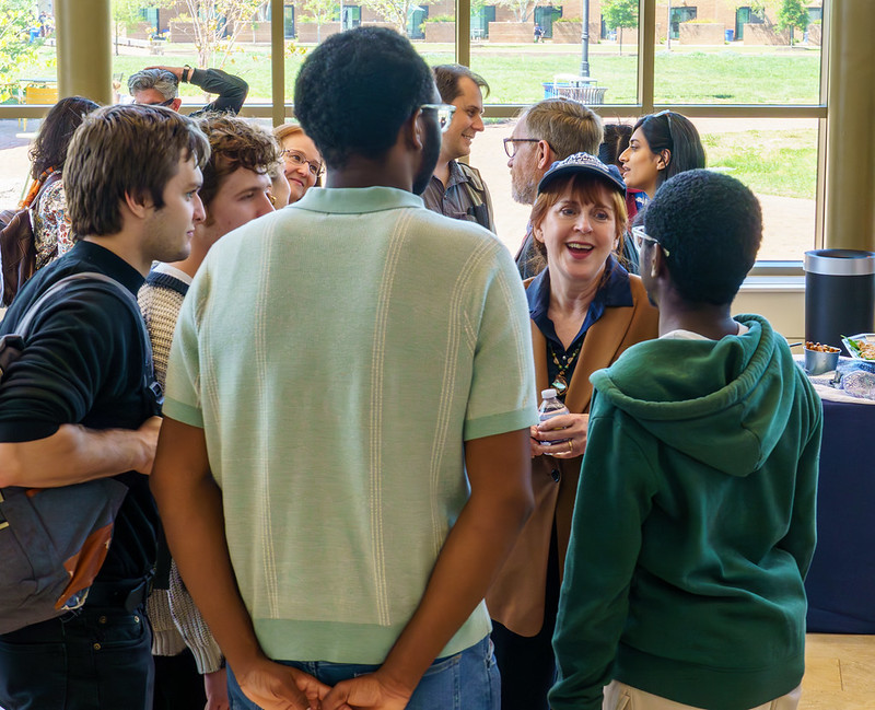 A group of people gathers indoors, conversing and smiling near large windows with a grassy outdoor view in the background.