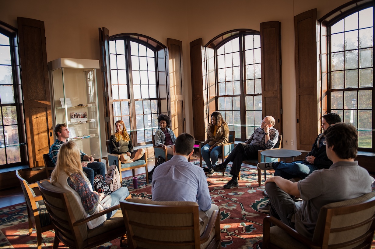 A group of people are seated in a circle having a discussion in a sunlit room with large windows and wooden chairs.