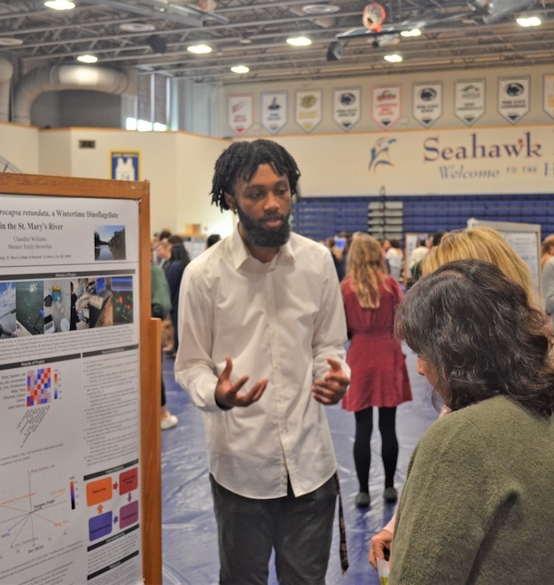 brochure8 A man in a white shirt discusses a research poster with a woman in a busy gymnasium.