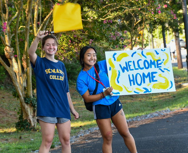 53939791537_4b54844697_c Two students outdoors, smiling and greeting. One waves a yellow flag, while the other holds a colorful sign that says "Welcome Home." They are near trees and bushes, conveying a sense of warmth and hospitality.
