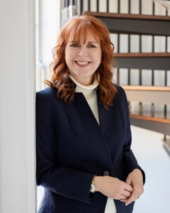 A woman with red hair wearing a navy blazer and white turtleneck stands indoors, smiling.