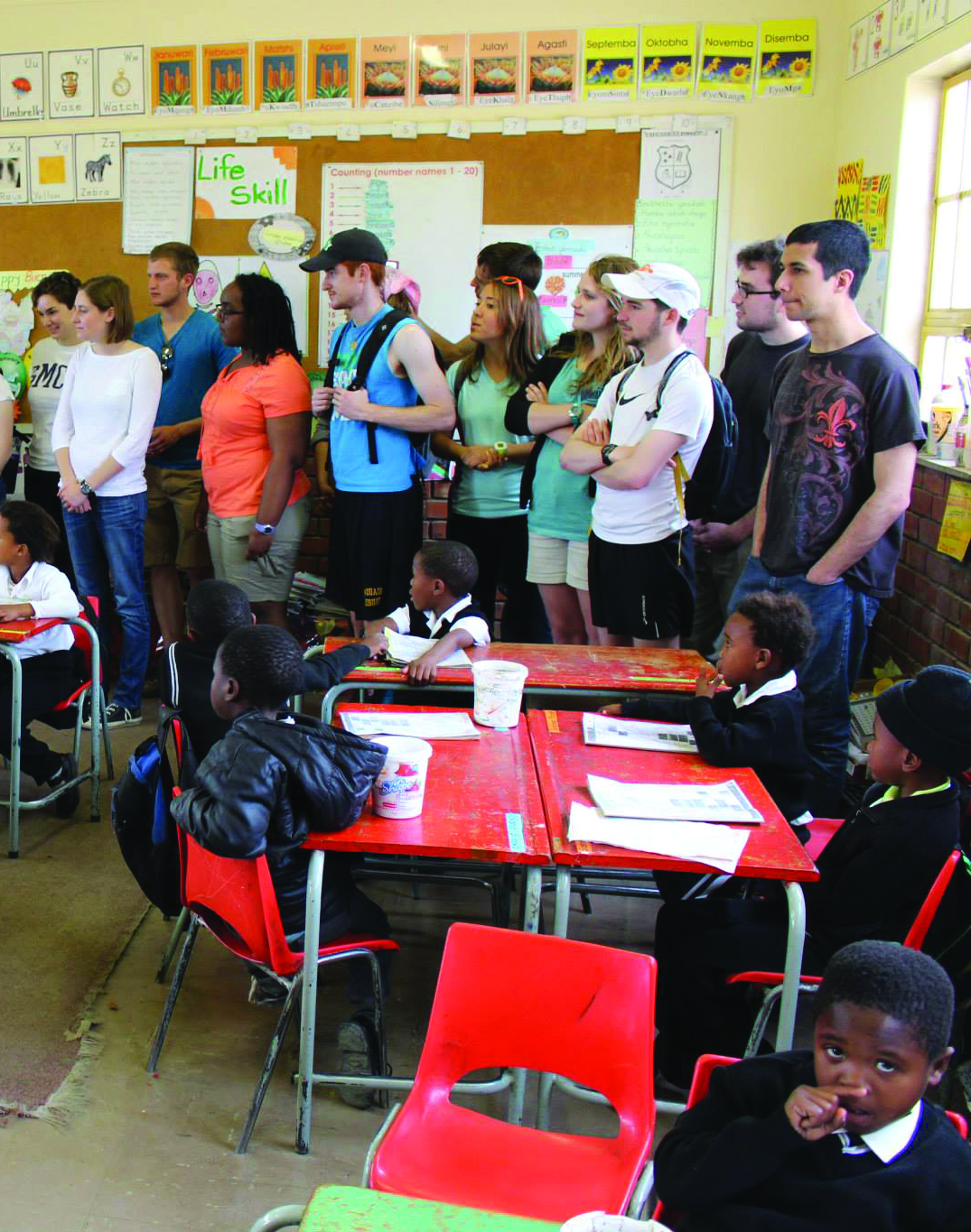 A group of international adults stands at the front of a classroom, observing while young students in uniform sit at desks and work on assignments.