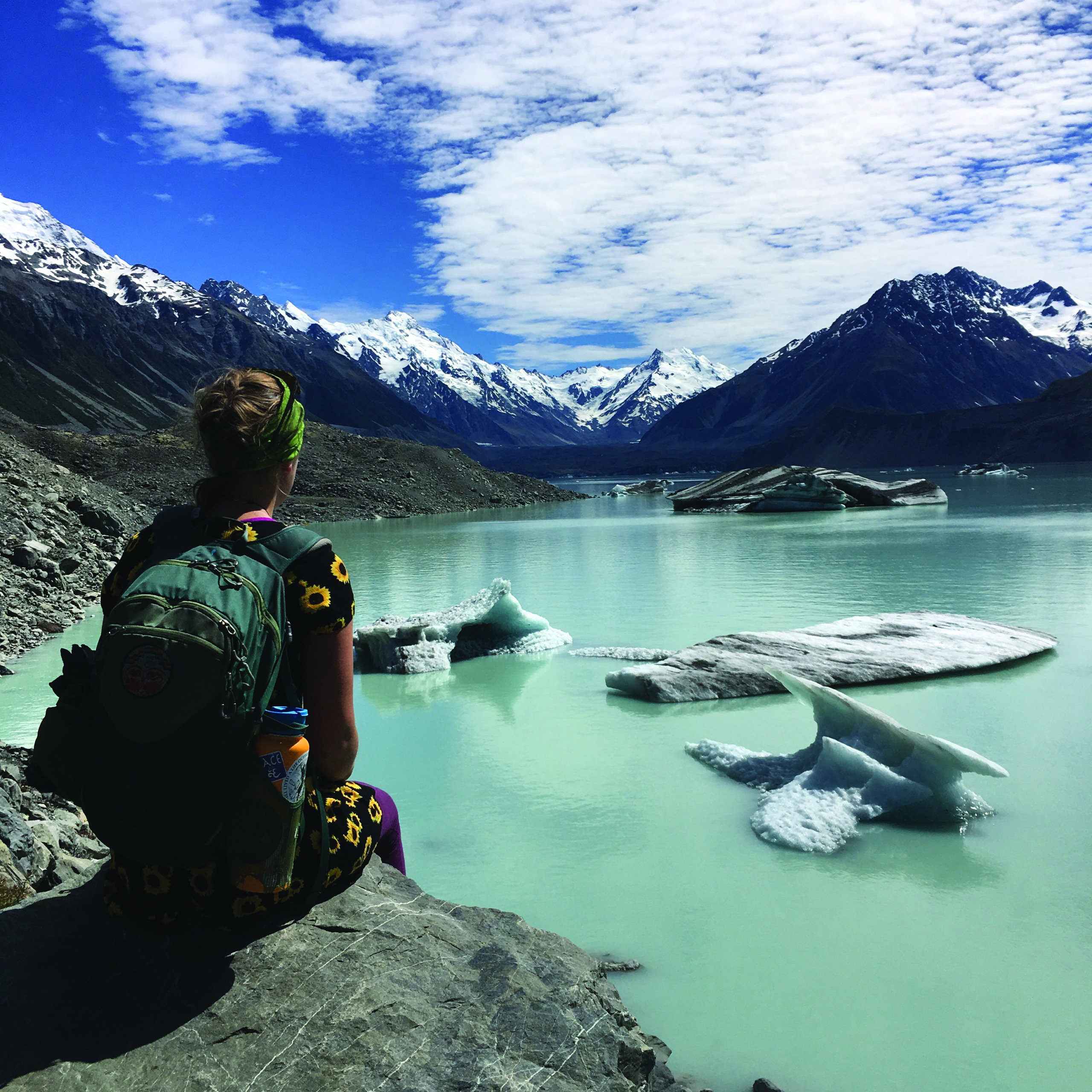 A person with a backpack sits on a rocky shore, gazing at icebergs floating in a glacial lake surrounded by international snow-capped mountains under a partly cloudy sky.