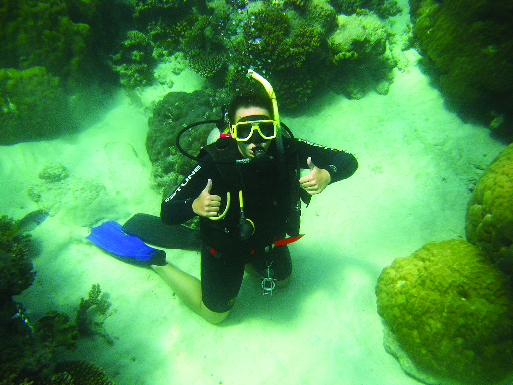 An international scuba diver kneels underwater on the sandy seabed, giving a thumbs-up gesture, surrounded by coral and rocks.