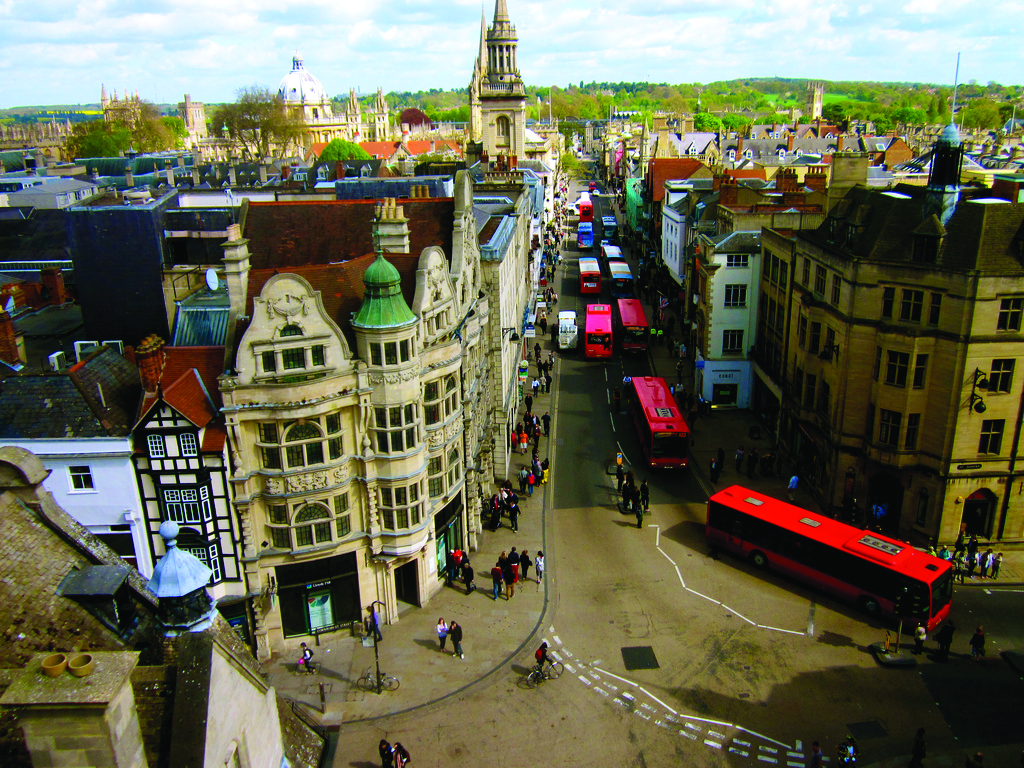 Aerial view of a busy city street with historic buildings, international red buses, pedestrians, and green hills in the background.