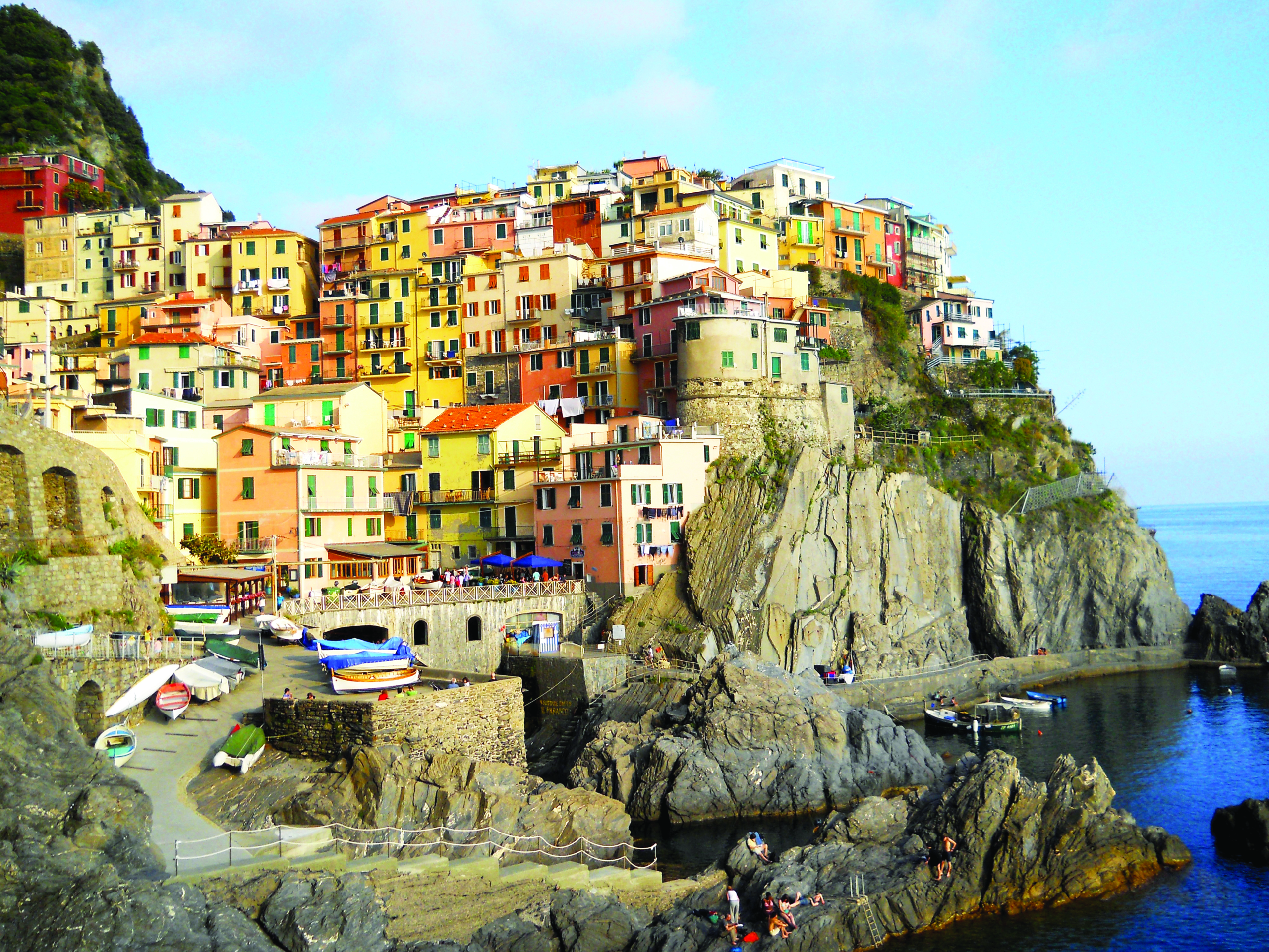 Colorful houses of Manarola, one of Cinque Terre’s international villages in Italy, perched on rocky cliffs above the Mediterranean Sea, with boats and people by the waterfront.
