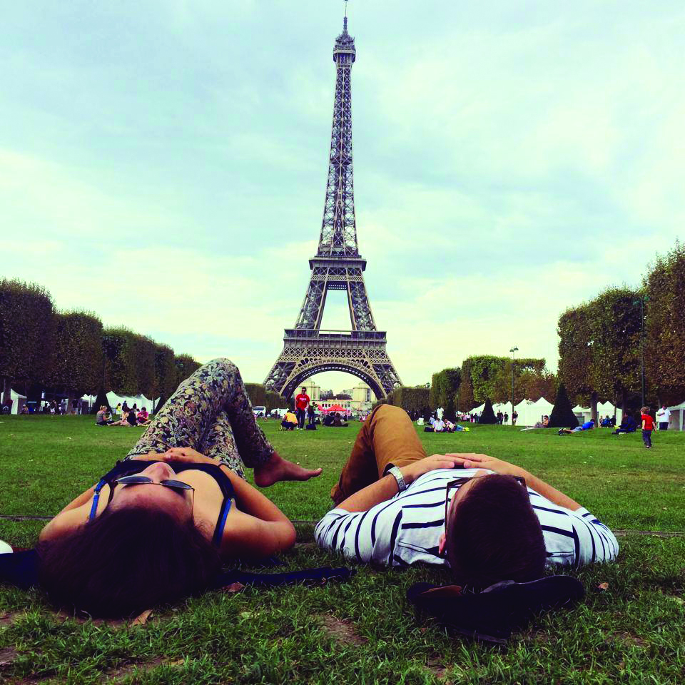 Two people lay on the grass facing the Eiffel Tower in Paris, enjoying an international atmosphere among visitors and trees on a cloudy day.