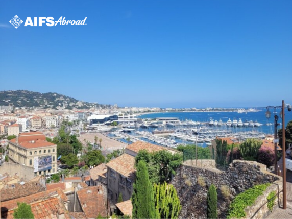 View overlooking a coastal city with a marina full of boats, red-tiled rooftops, hills in the background, and clear blue sky; "AIFS Abroad" logo in the top left corner.