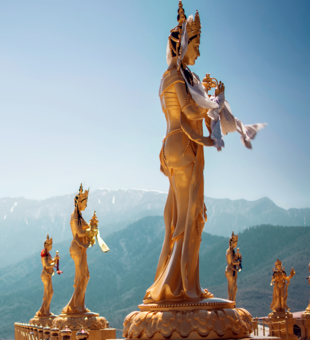Golden statues of deities stand on a platform overlooking a range of green mountains under a clear blue sky.