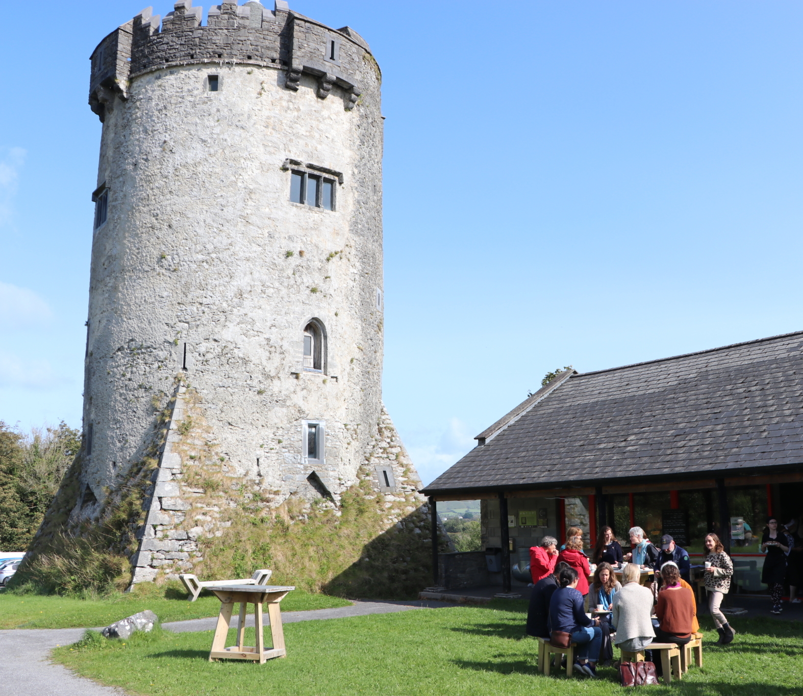 A group of people sit and stand around picnic tables outside a stone tower and modern building on a sunny day.