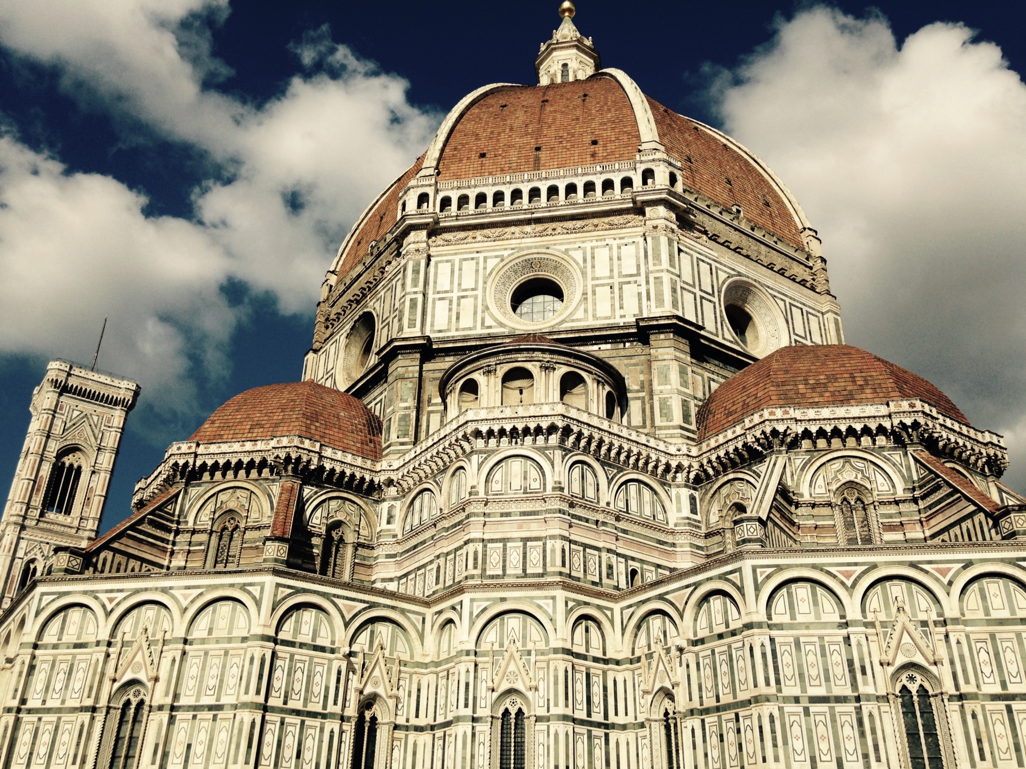 The image shows the exterior of Florence Cathedral with its large red-tiled dome, ornate marble façade, and a partly visible bell tower against a cloudy sky.