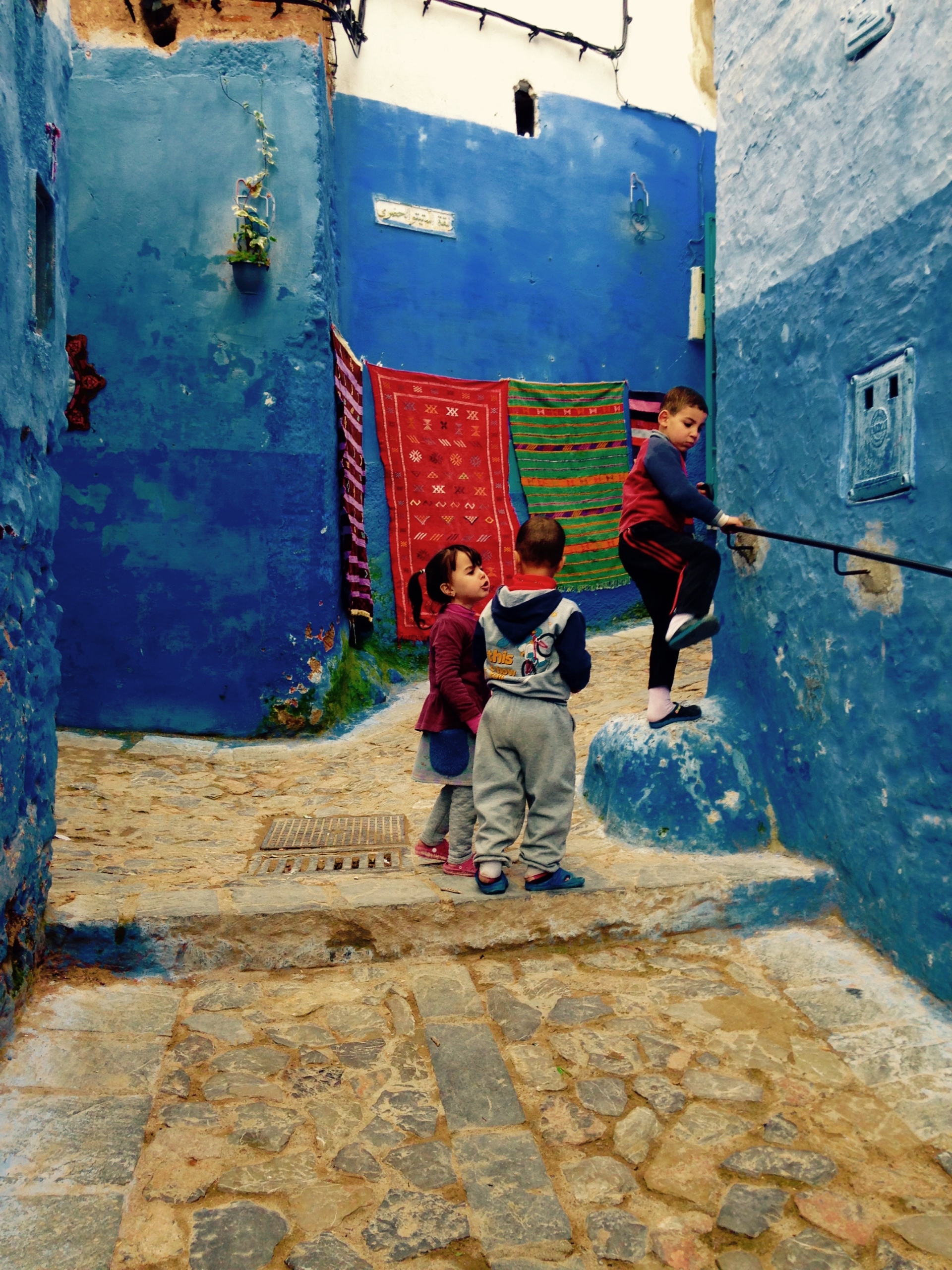 Three children play on a narrow stone street with blue-painted walls and colorful rugs hanging in the background.